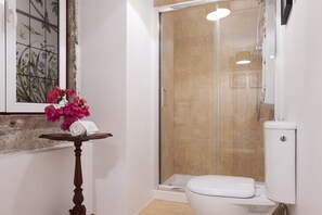 Un baño con ducha de vidrio, inodoro y una mesita con una toalla enrollada y flores rosadas.