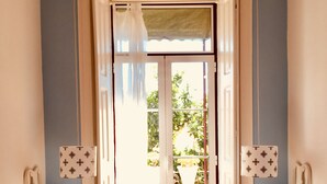 Quarto com duas camas, cada uma com travesseiro e mesa de cabeceira. Há uma janela com cortinas e vista para o verde lá fora.
