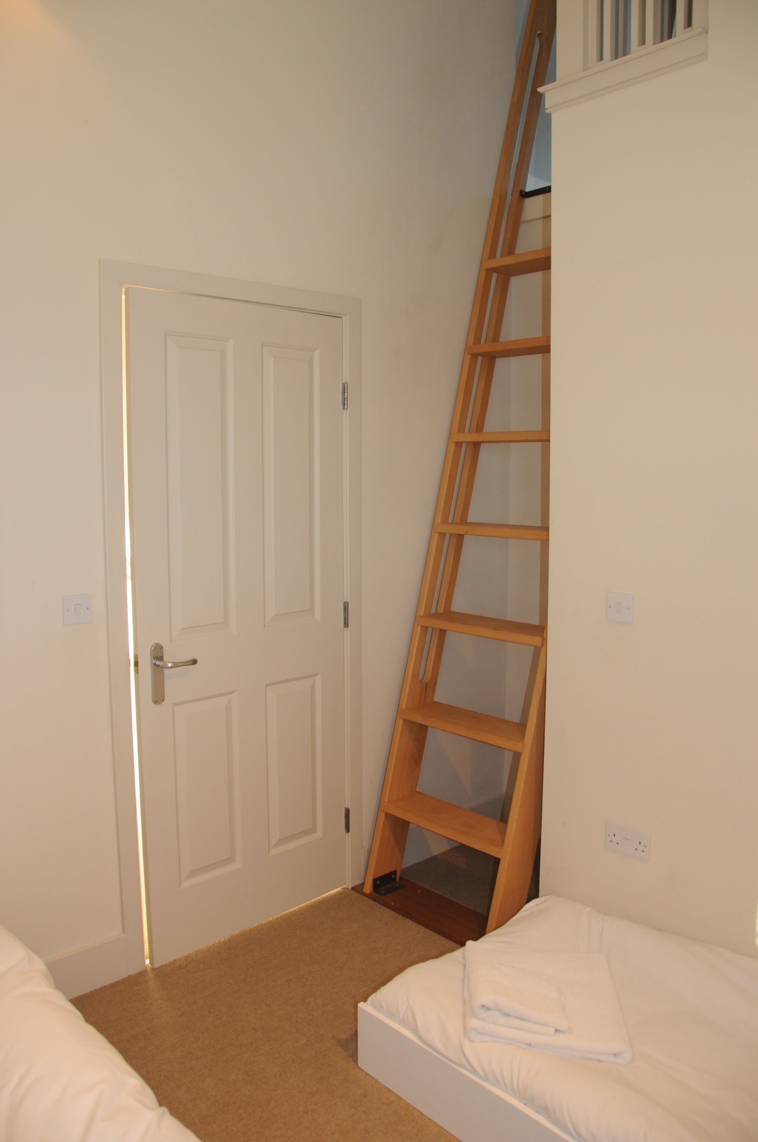 A bedroom with a white door, a wooden ladder, and a bed with white bedding.