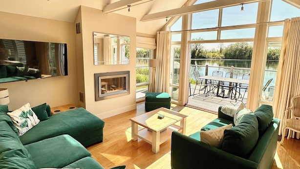 A spacious living room with a large green sofa set, a wooden coffee table, and a fireplace. There is a large window offering a view of a body of water and outdoor seating. - Beautiful 4 bed lakeside lodge in the Cotswolds. Private deck & STUNNING views (South Cerney)
