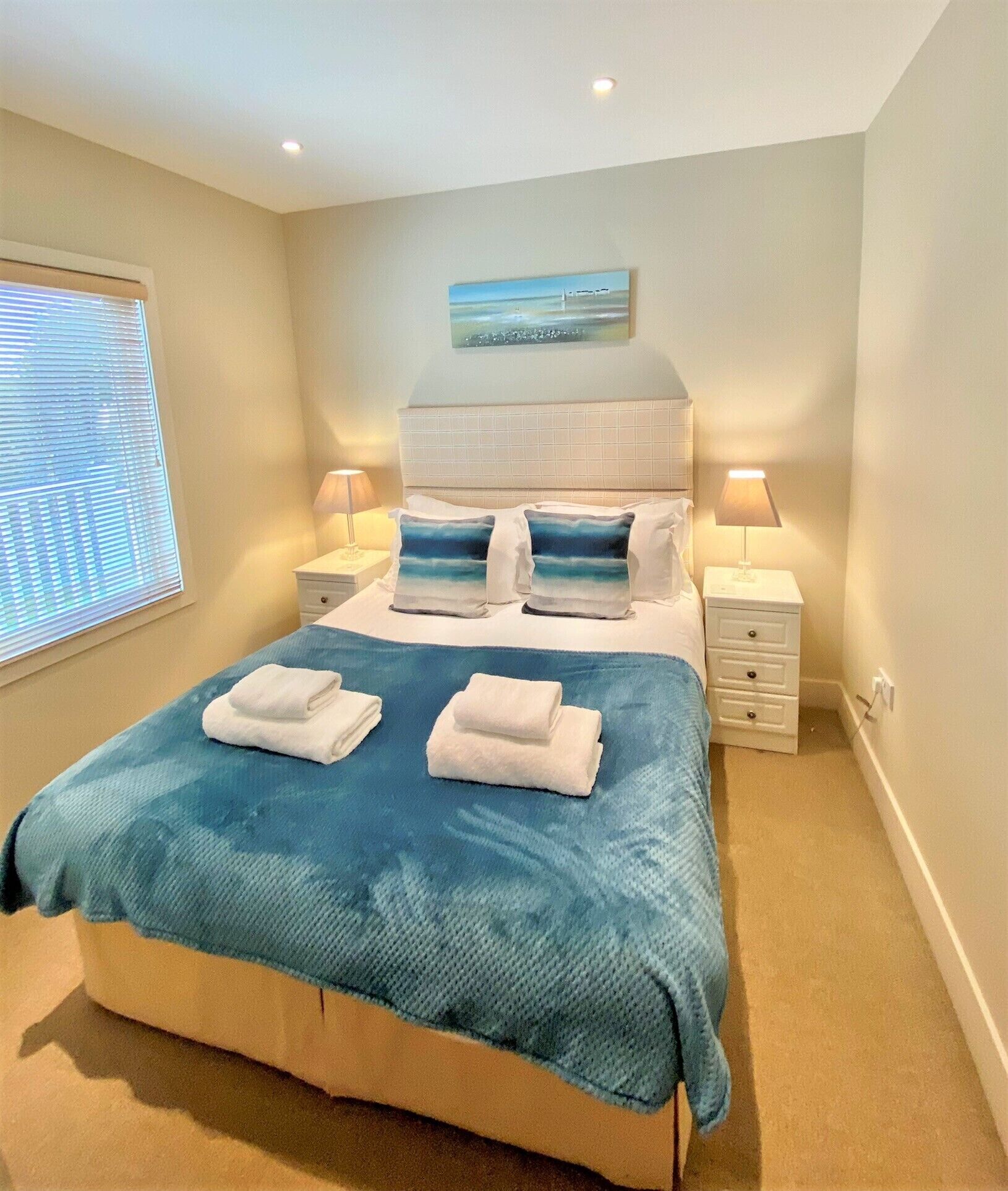 A neatly made bed with a blue blanket, two pillows, and folded towels. There is a nightstand with a lamp, a window with blinds, and a painting on the wall above the bed.