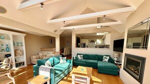 A spacious living room with a green sectional sofa, a white armchair, and a wooden coffee table. A fireplace is visible on the right, and a kitchen area is in the background. - Beautiful 4 bed lakeside lodge in the Cotswolds. Private deck & STUNNING views (South Cerney)