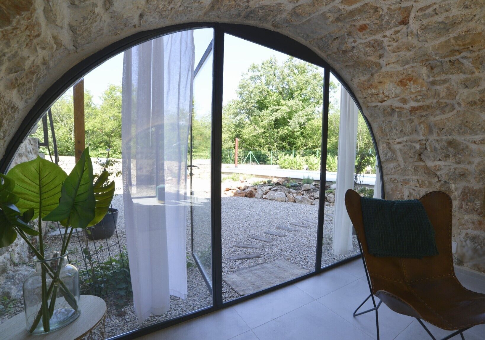 A room with a stone wall, a large arched window, a chair, and a potted plant.