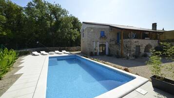 A swimming pool with lounge chairs, a stone building with a tiled roof, and surrounding greenery.