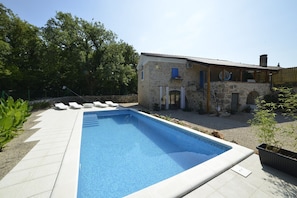 Piscina com espreguiçadeiras, um edifício de pedra com telhado de azulejos e vegetação ao redor.