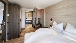 A modern hotel room with a wooden floor, a large bed with white linens, a glass partition, and a bathroom visible through the partition. - Panorama Living Dolomites (Rio di Pusteria)