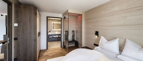 A modern hotel room with a wooden floor, a large bed with white linens, a glass partition, and a bathroom visible through the partition.