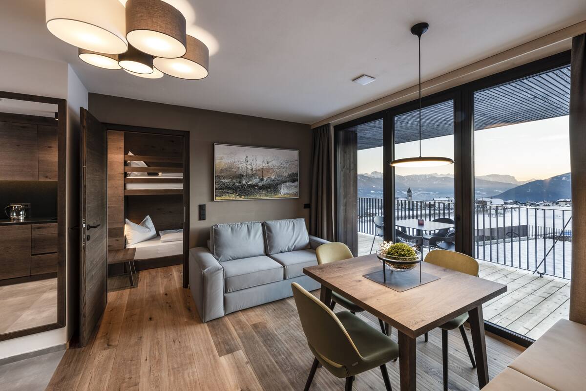 A modern hotel room with a balcony, a sofa, a dining table, and a large window offering a mountain view.
