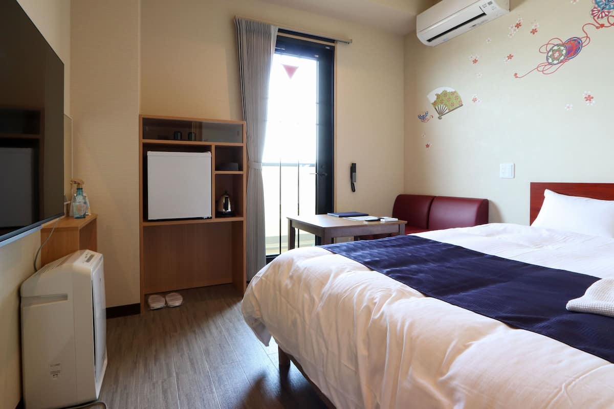 A hotel room with a bed, a desk, a chair, a wardrobe, a mirror, an air conditioner, and a window with curtains.