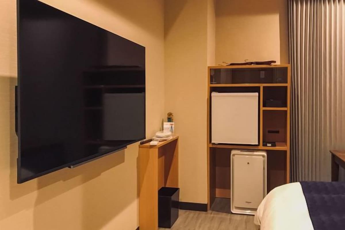 Habitación de hotel con televisor de pantalla plana, un mini refrigerador, una pizarra y un estante de madera.