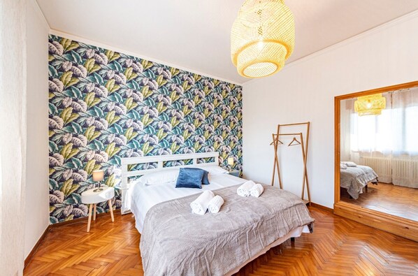 Une chambre avec un papier peint à motifs, un lit avec une tête de lit blanche, une table de chevet avec une lampe et un plancher en bois.