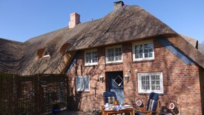 Ein reetgedecktes Cottage mit Ziegelaußenwand, ein kleiner Garten mit Topfpflanzen und ein Tisch für zwei Personen mit Stühlen.