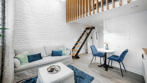 Una sala de estar moderna con pared de ladrillo blanco, una escalera de madera, un sofá con cojines, una mesa de centro con mandos a distancia y dos sillones alrededor de una mesita con un jarrón.