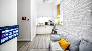 Un espacio habitable moderno con una televisión de pantalla plana, un sofá con un cojín amarillo, una zona de cocina con encimera de madera y una pared de ladrillo blanco.