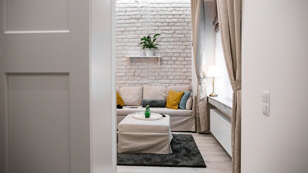 Una sala moderna con pared de ladrillo blanco, un sofá color beige con cojines amarillos, un estante blanco con una planta y un tapete negro.