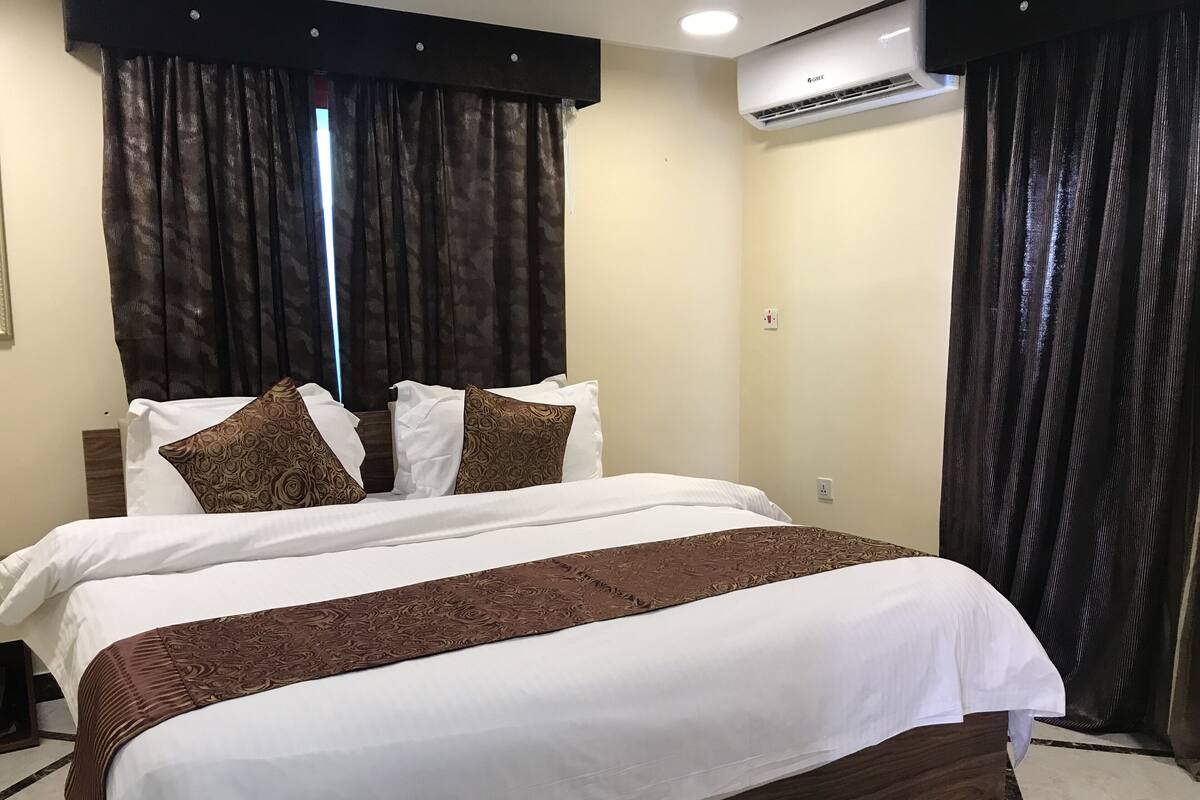 A hotel room with a large bed, dark curtains, an air conditioning unit, and a tiled floor.
