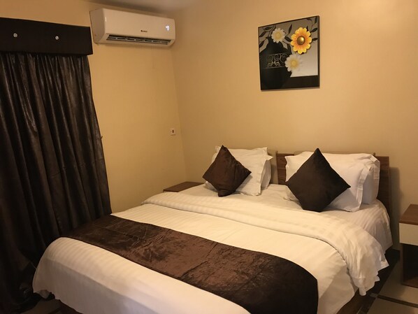 A hotel room with a bed, a nightstand, a wall-mounted air conditioner, and a painting of flowers.