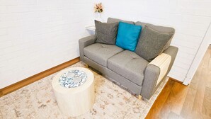 Harmaa sohva, jossa on sininen koristetyyny, pyöreä puinen sivupöytä ja kuviollinen matto huoneessa, jonka seinät ovat valkoista tiiltä.