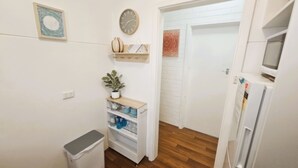 Pieni, siisti huone, jossa on valkoinen ovi, kello, koriste-esineitä sisältävä hylly, ruukkukasvi ja roskakori.