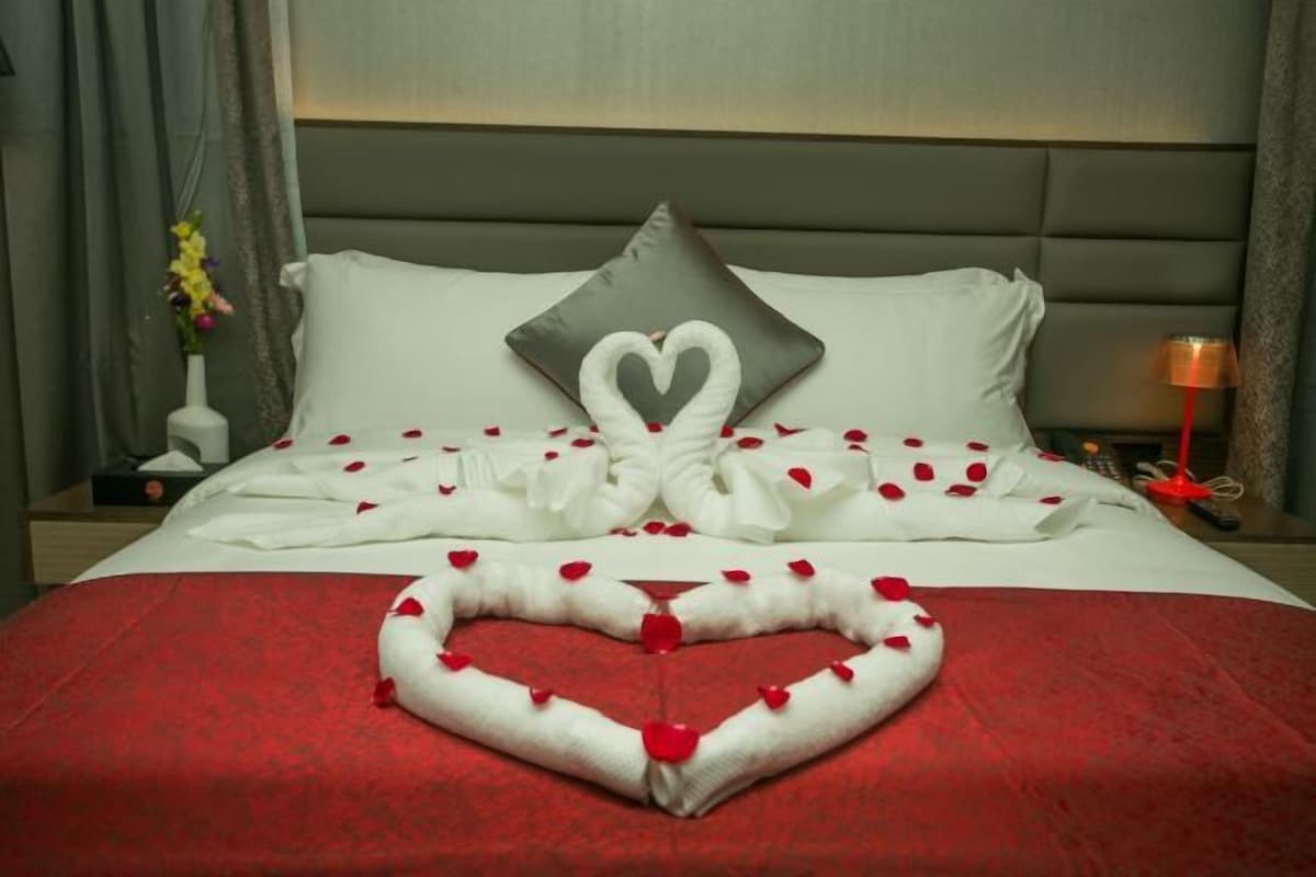 A hotel room with a bed, pillows, a heart-shaped decoration made of rose petals, and a vase with flowers.