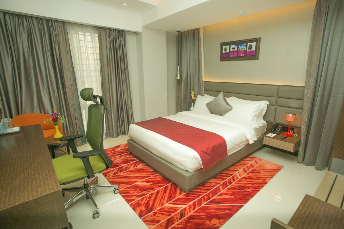 A modern hotel room with a large bed, a desk, a chair, and a nightstand with a lamp.