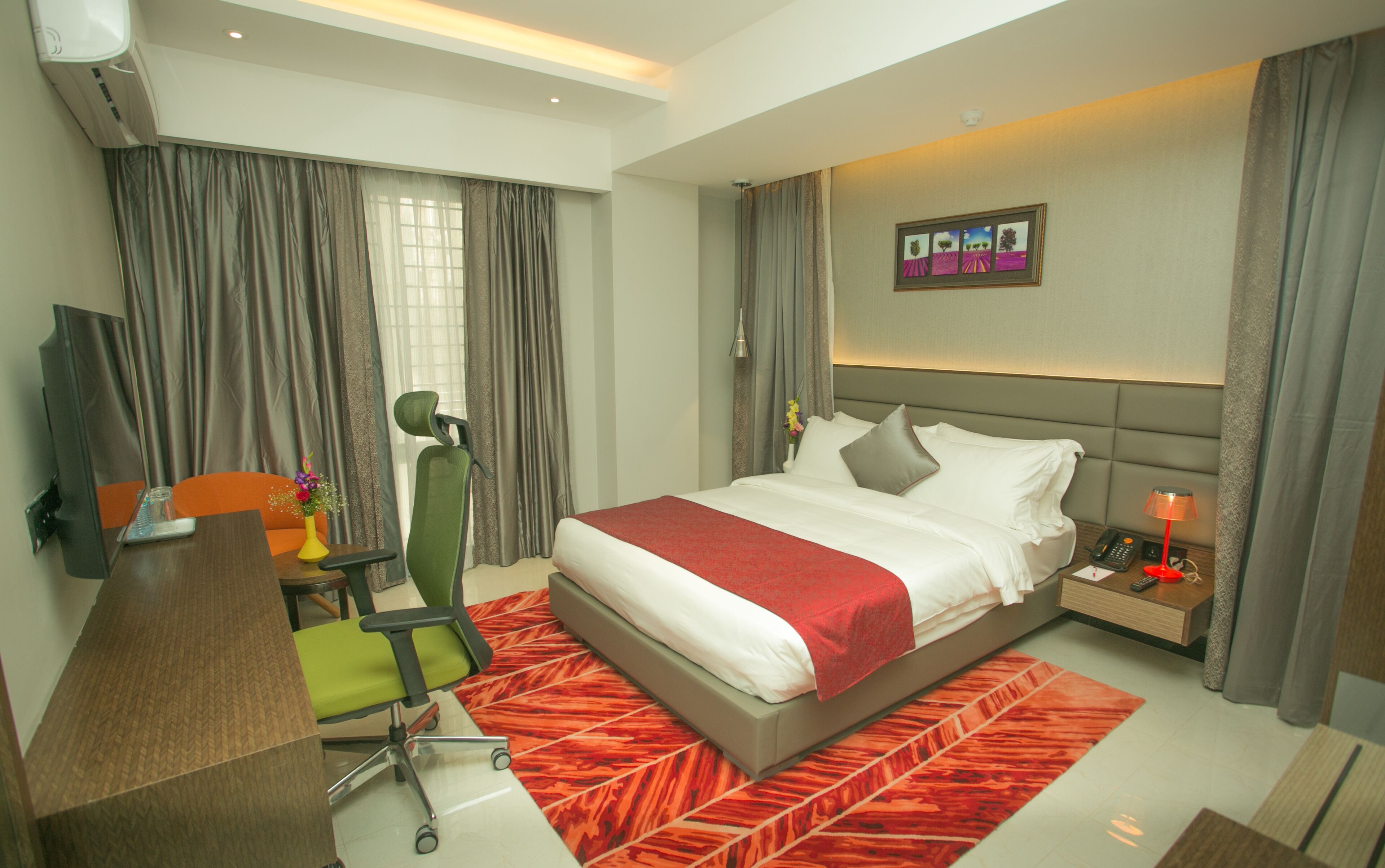 A modern hotel room with a large bed, a desk with a chair, a TV, and a bedside table with a lamp.