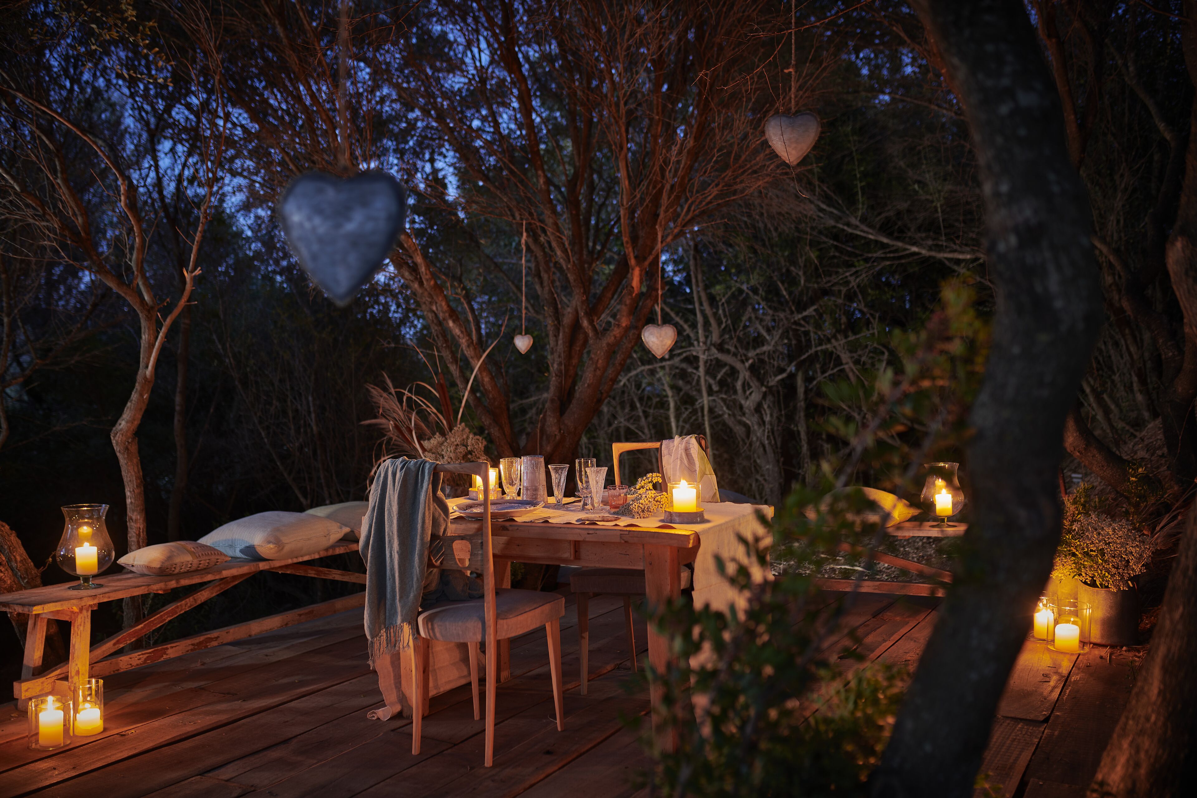 An outdoor dining setup with a wooden table, chairs, candles, and heart-shaped decorations.