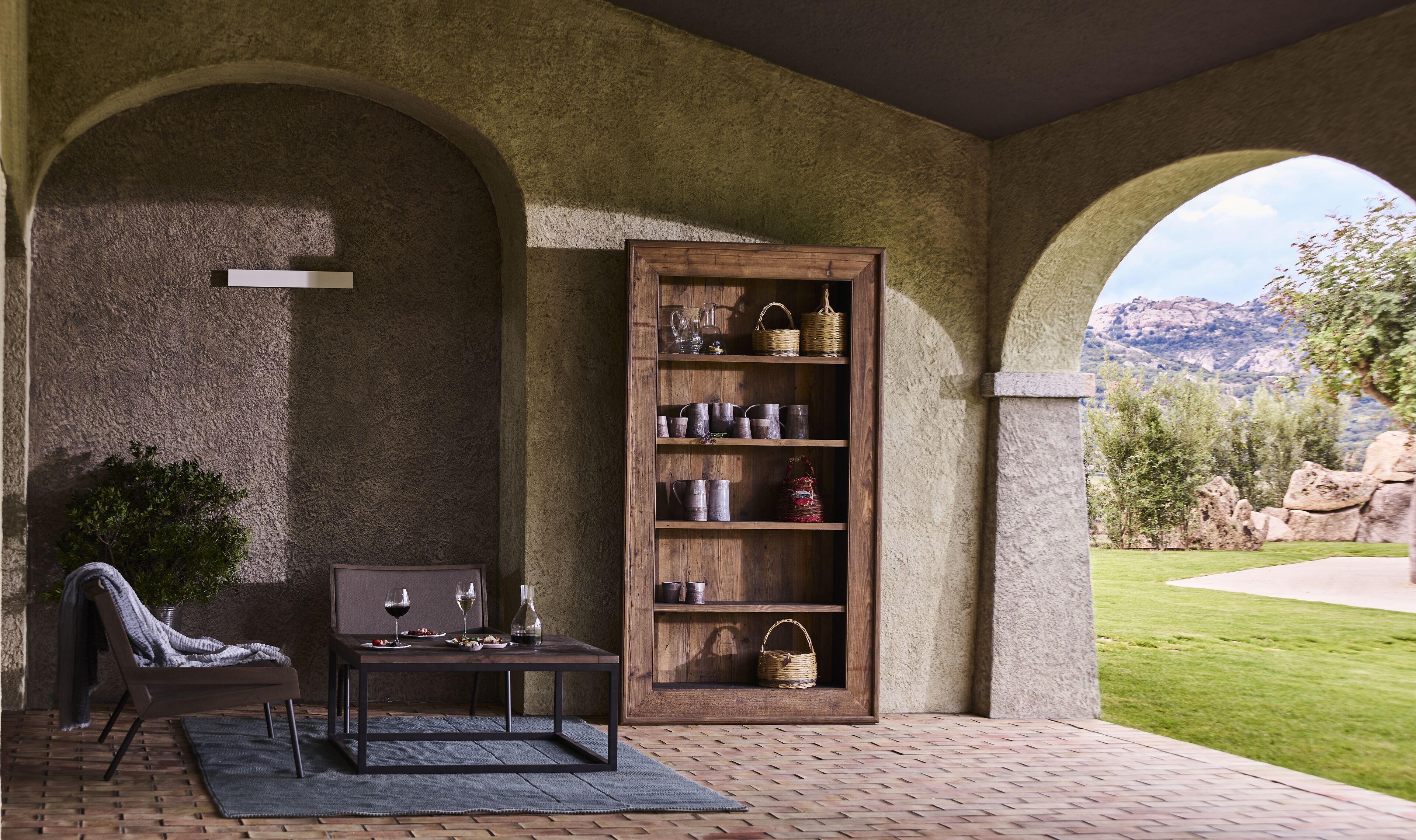 A cozy outdoor seating area with a table and chairs, a wooden bookshelf, and a view of a mountainous landscape.