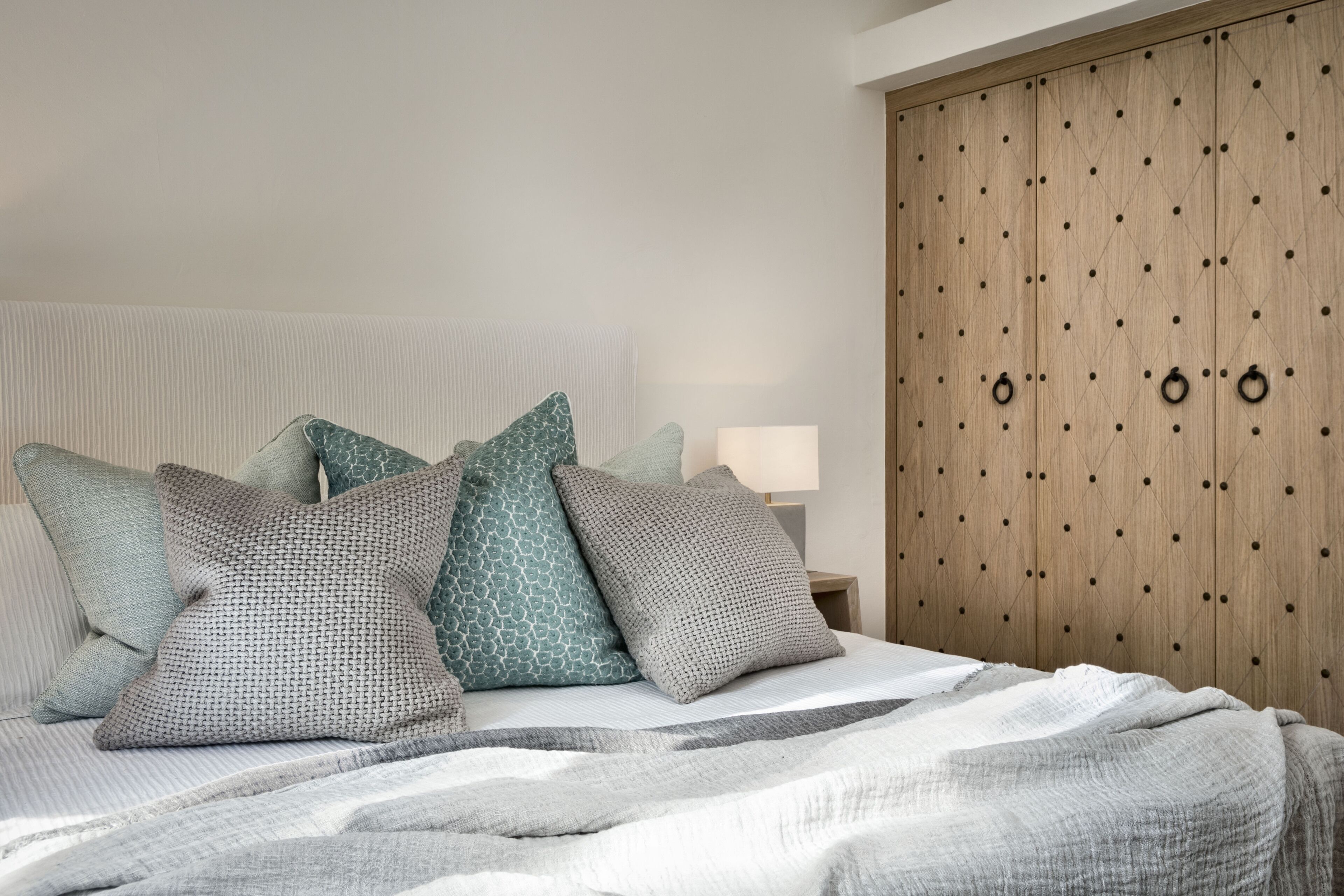 Uma cama bem arrumada com manta listrada, várias almofadas e um guarda-roupa de madeira com puxadores redondos.