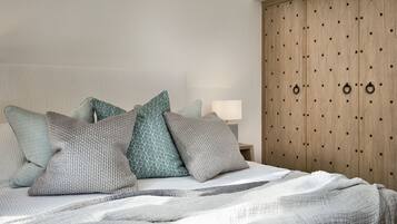 Uma cama bem arrumada com manta listrada, várias almofadas e um guarda-roupa de madeira com puxadores redondos.
