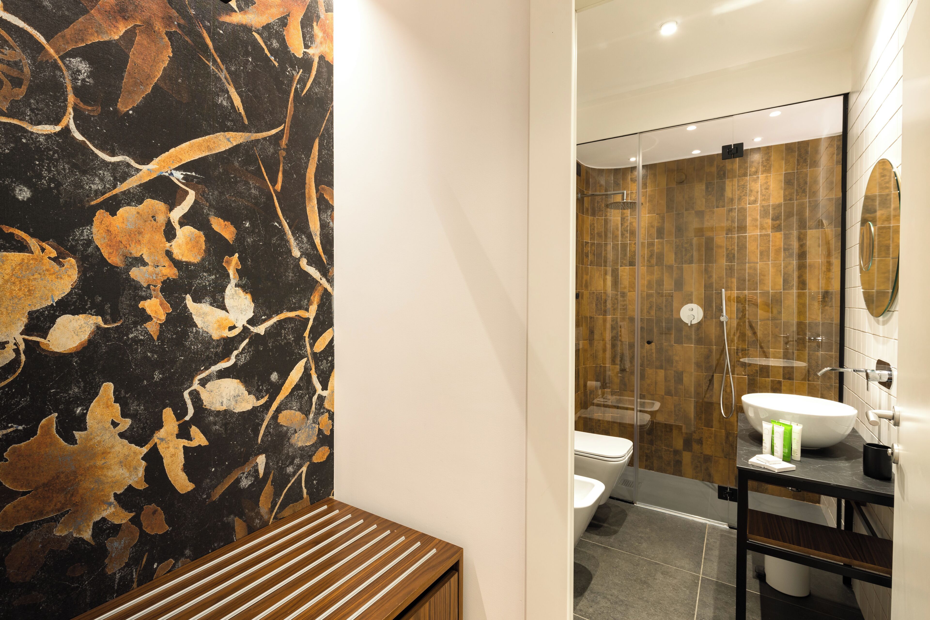 A modern bathroom with a black and gold marble wall, a wooden bench, a glass-enclosed shower, and a black vanity with a white sink.