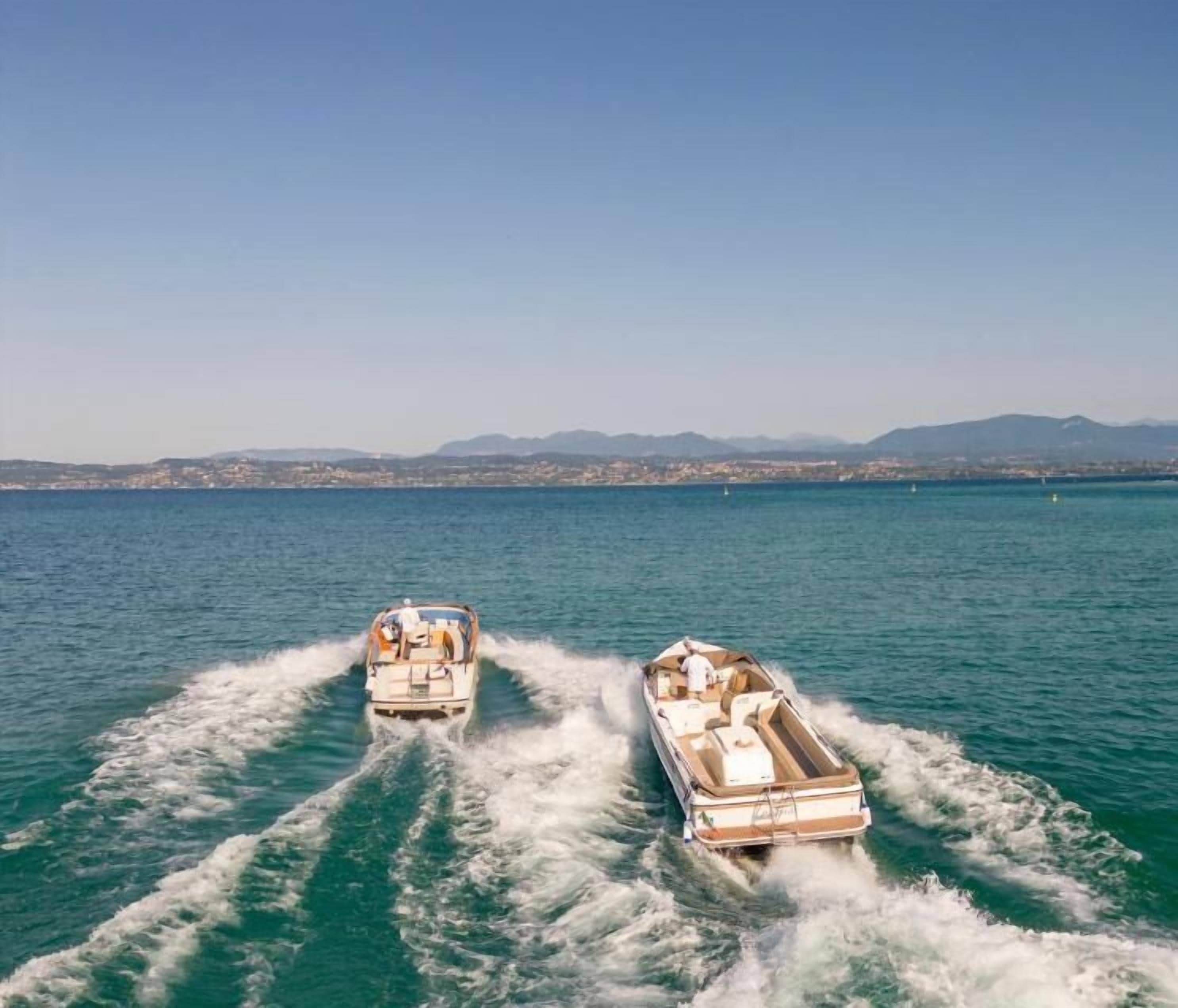Two boats moving through a body of water with a distant shoreline and mountains in the background.