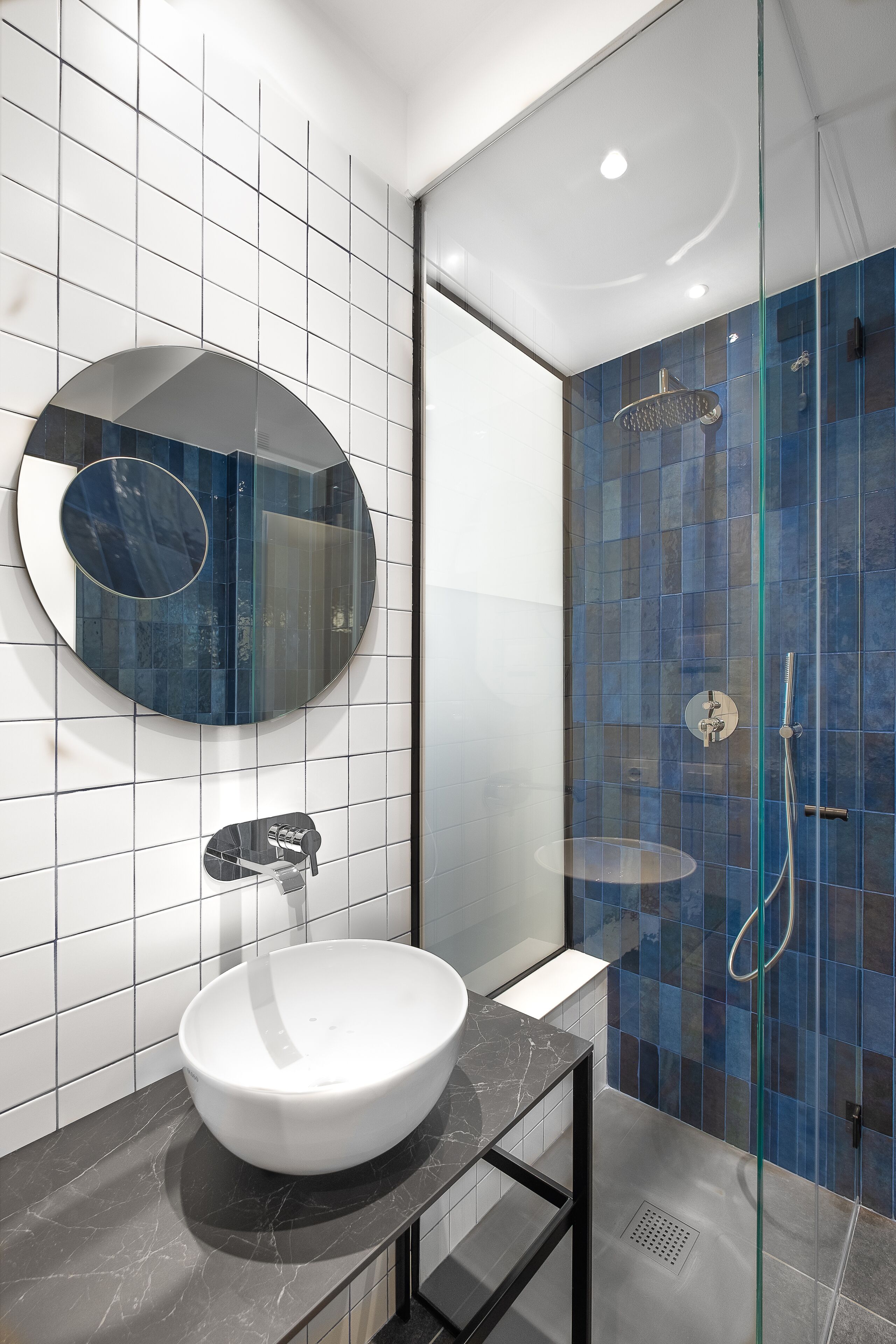 A modern bathroom with a white sink, a round mirror, and a glass-enclosed shower area with blue tiles.