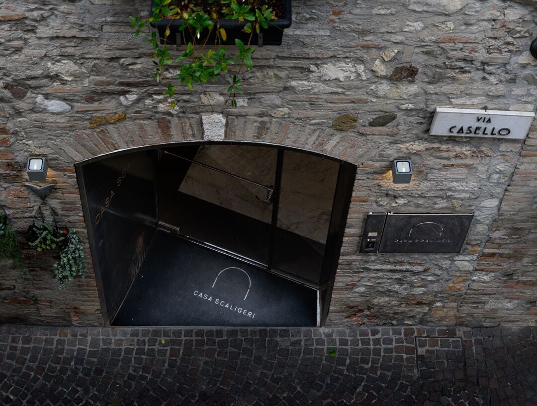 A stone wall with a black metal structure, a sign reading "VIA CASELLO," and a plaque with "CASA SCALIGERI" text.