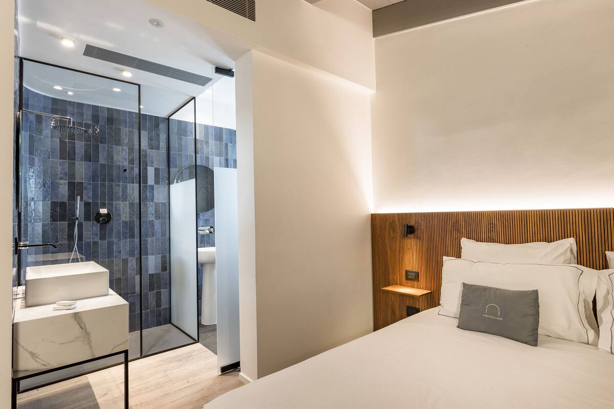 Una habitación de hotel moderna con ducha de vidrio, lavamanos de mármol, cabecero de madera y una cama bien hecha con un almohadón.