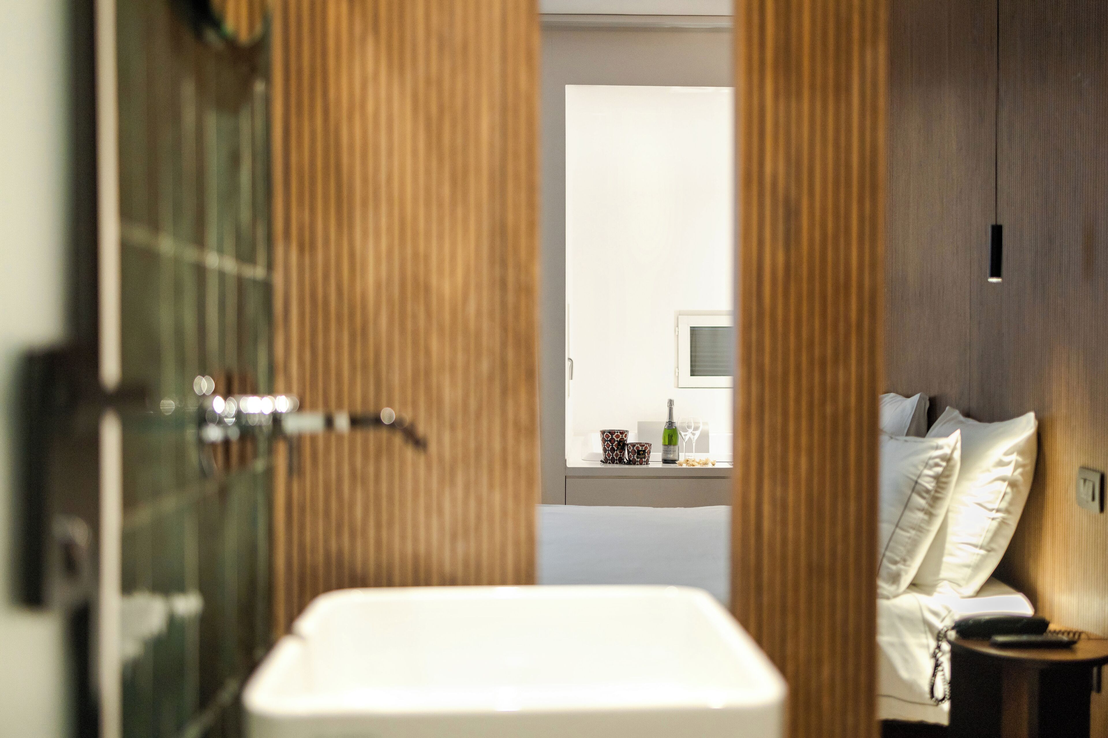 A hotel room with a bathroom, a bed, a bedside table, and a small table with a bottle and glasses.