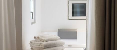 A hotel room with a bed, a window with blinds, a small table with folded towels, and a wall-mounted air conditioning unit.