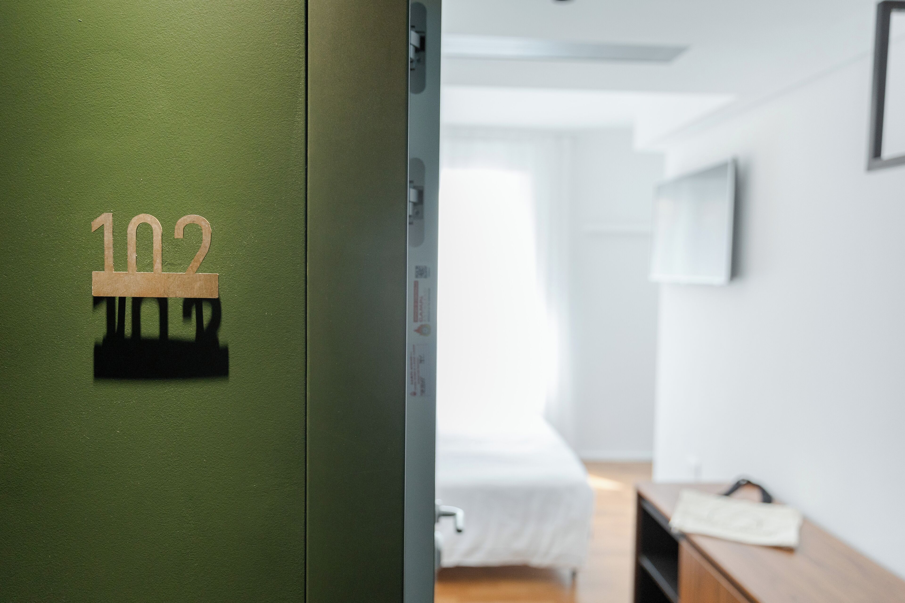 Habitación de hotel con una puerta verde marcada '102', una cama, una mesita de noche y una ventana.