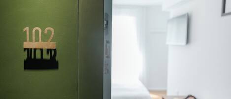Habitación de hotel con una puerta verde marcada '102', una cama, una mesita de noche y una ventana.