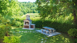 A stone barbecue area with a table and benches, surrounded by lush greenery and a small pond. - Wonderful new villa, lush fenced garden, private pool (Pazin)