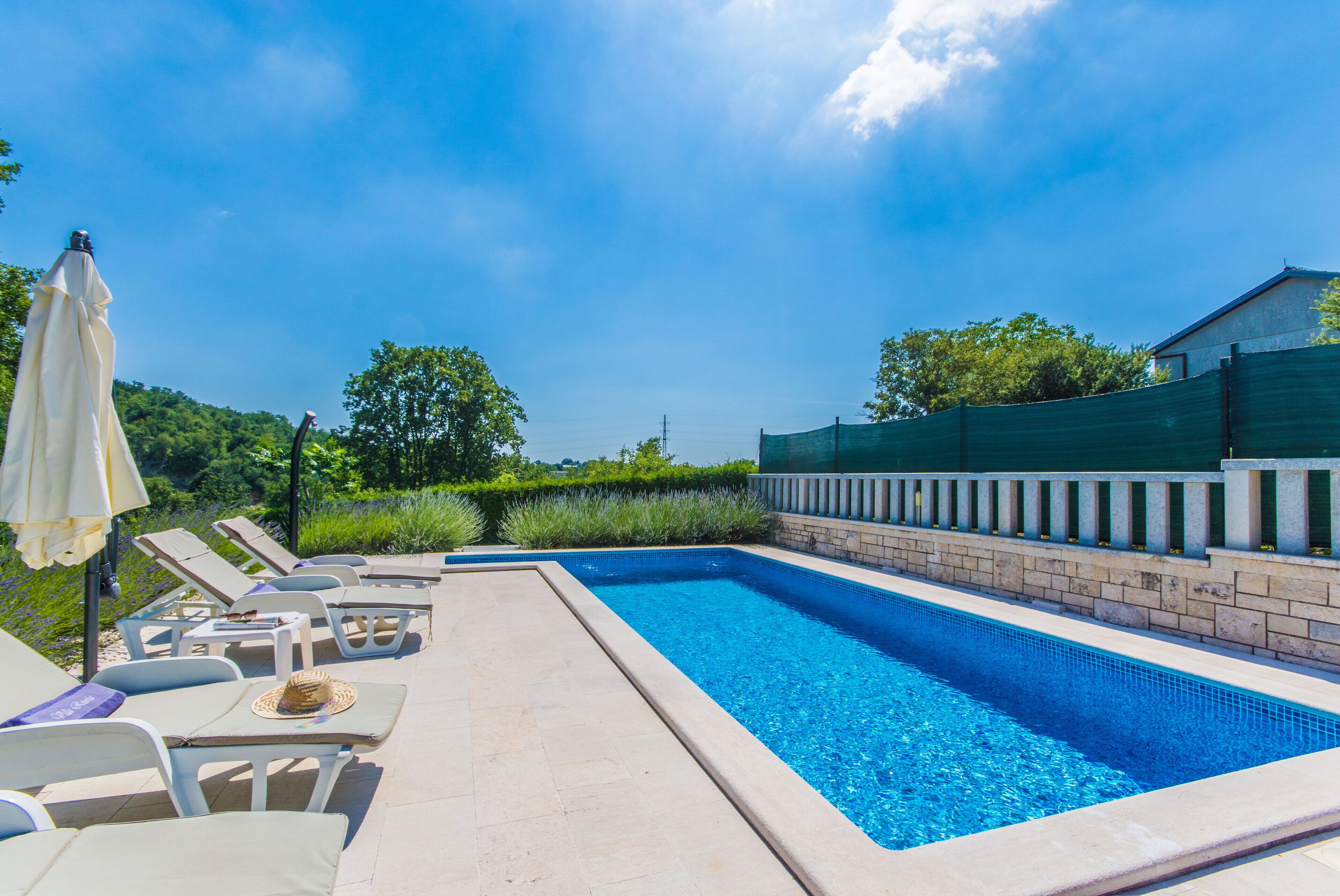 Ein rechteckiger Swimmingpool mit Fliesenrand, umgeben von Liegestühlen und einem Tisch mit Sonnenschirm.