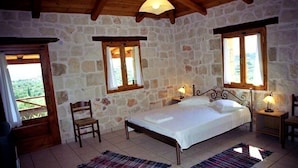 Un dormitorio con pared de piedra, una cama, dos ventanas, una silla y techo de madera.