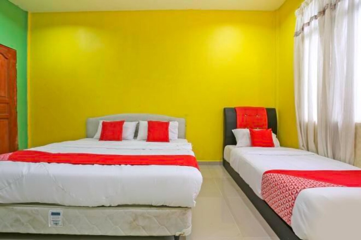 A room with two beds, each with white bedding and red accents. The walls are painted yellow, and there is a wooden door on the left.