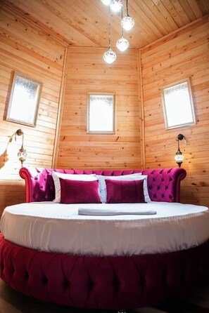 Ein rundes Bett mit einer roten, gepolsterten Kopfteil in einem Holzraum mit Lamellenwänden und zwei Fenstern.