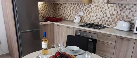 A modern kitchen with wooden cabinets, a round dining table set for two, and a breakfast setup including a bowl of fruit, a bottle of wine, and a candle.