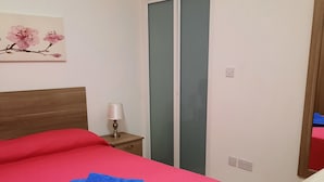 A bedroom with a wooden headboard, a red bedspread, a blue towel, a bedside lamp, and a wall-mounted air conditioner. - Porto Paolo side sea view ( Condominium ) (St. Paul's Bay)