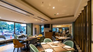 Un restaurante moderno con mesas de madera y sillas verdes, amplias ventanas que dan a la calle y un televisor montado en la pared.