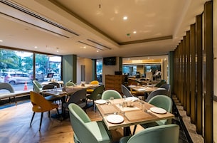 A modern restaurant with wooden tables and green chairs, large windows with a view of the street, and a TV mounted on the wall.