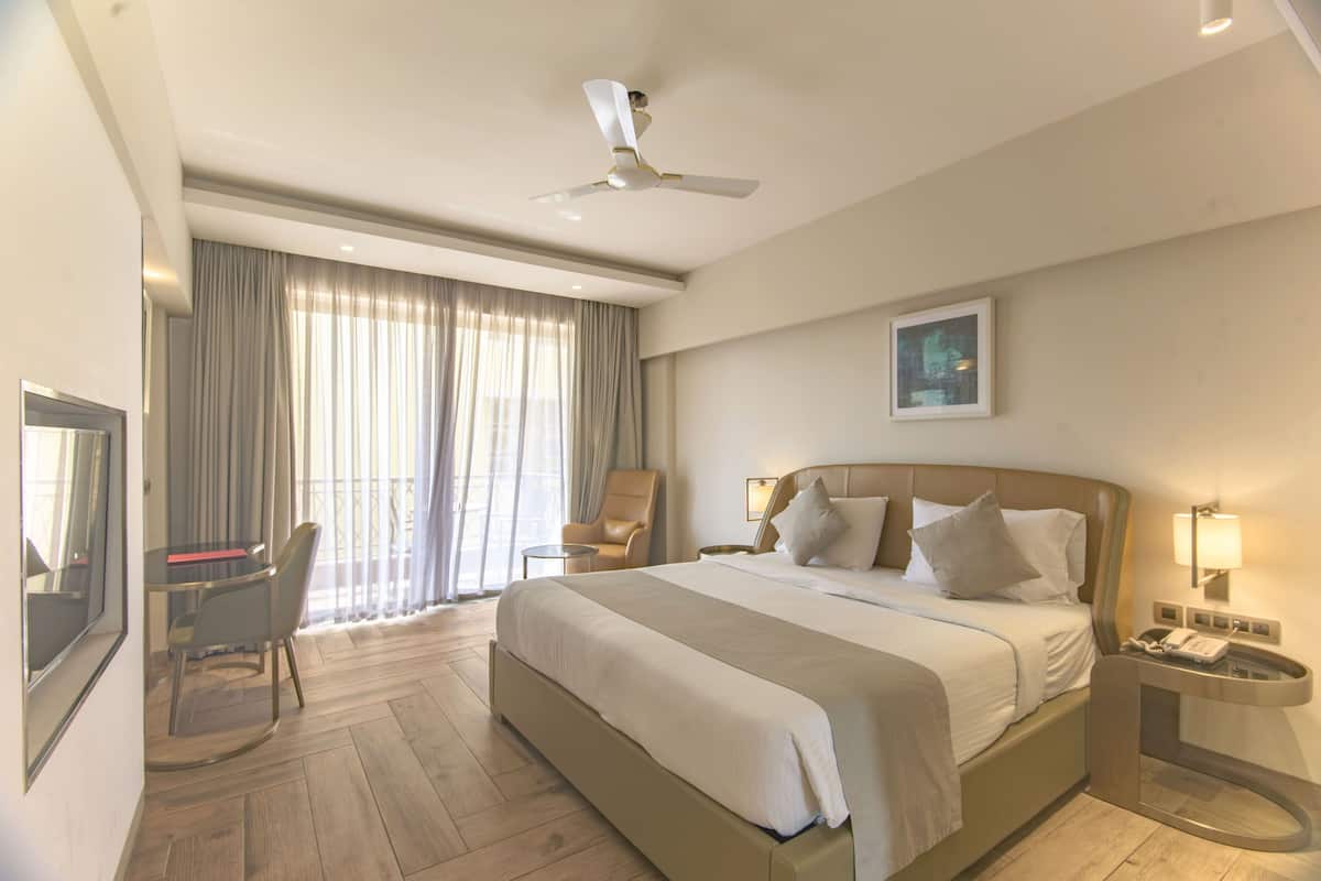 A hotel room with a large bed, a desk with a chair, a TV, a ceiling fan, and a window with curtains.