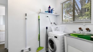 A laundry room with a washing machine, a broom, a mop, and a window with blinds. - Beauty AND THE Peace - Inlet Side (Inverloch)